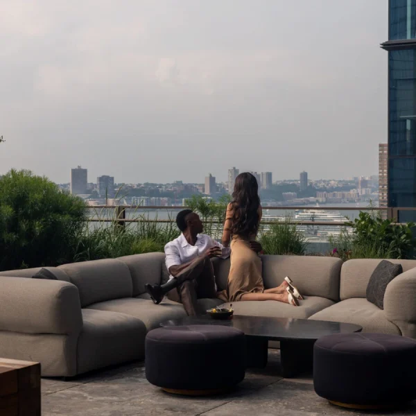 Two people on curved rooftop sofa with coffee table, greenery, and riverfront city view in background