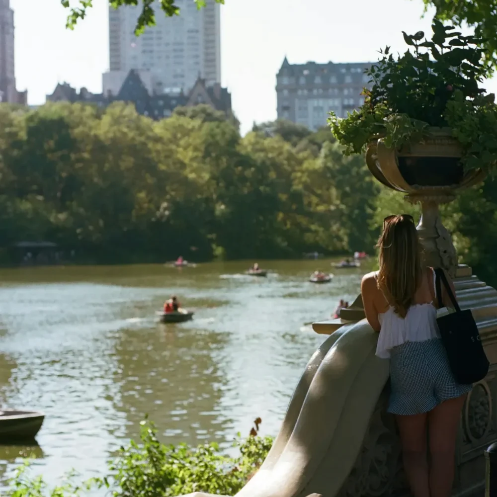 Person standing on a stone railing overlooking a lake with rowboats in Central Park, surrounded by trees & city buildings.