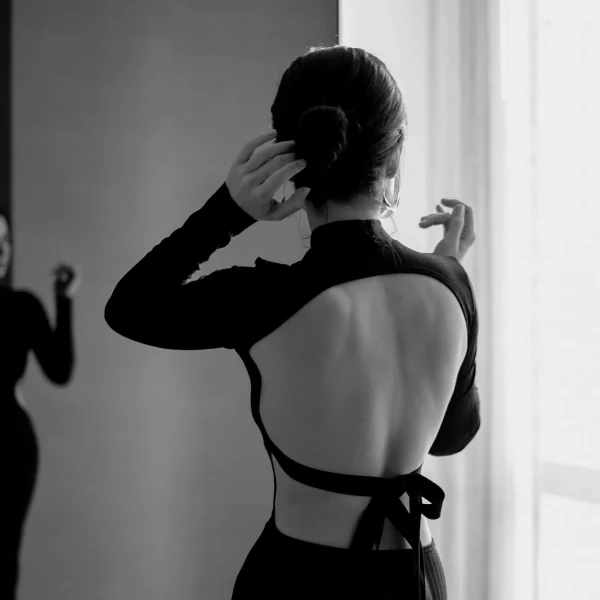 Woman in open-back dress adjusting hair in front of a mirror with soft natural light.