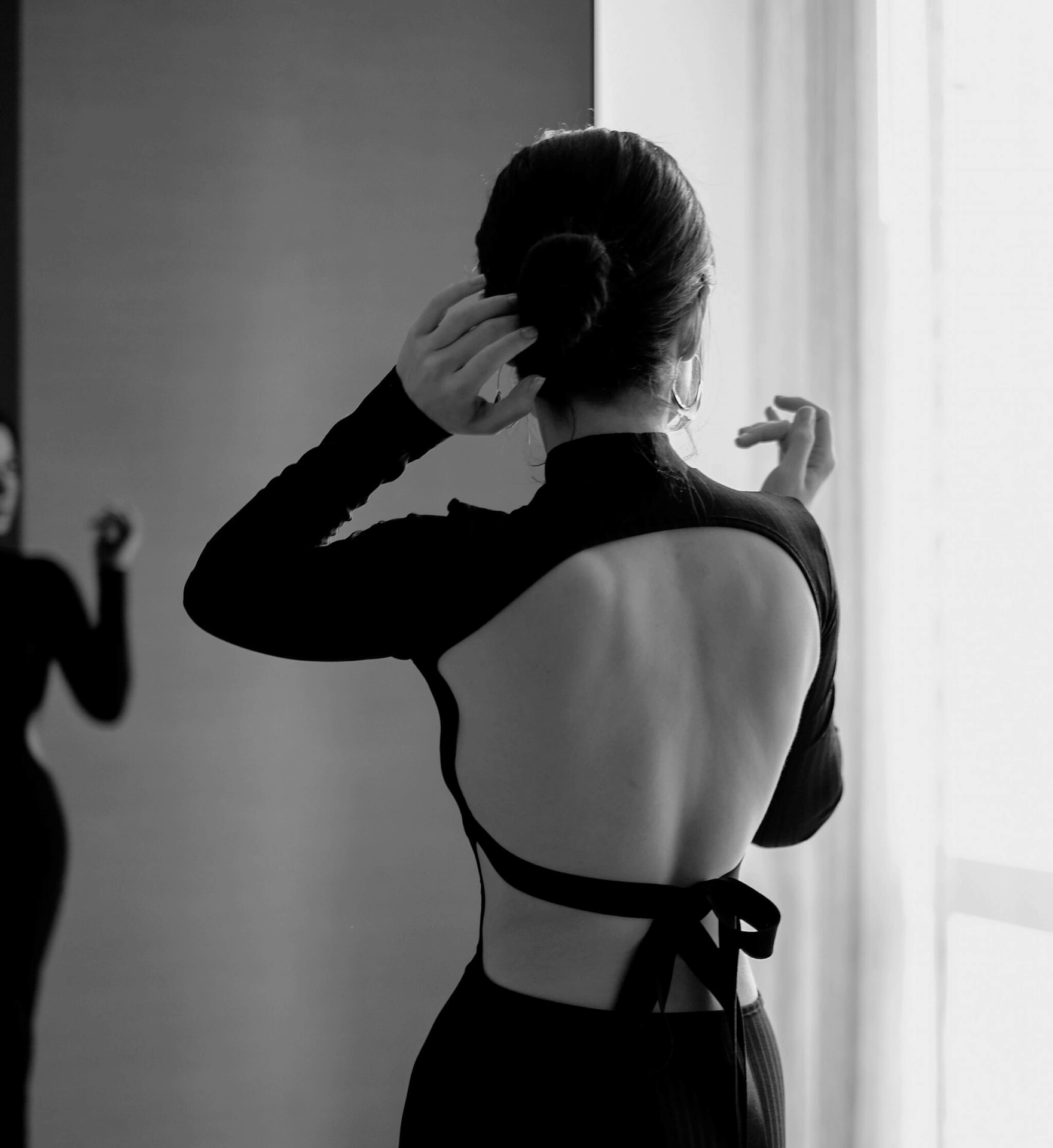 Woman in open-back dress adjusting hair in front of a mirror with soft natural light.