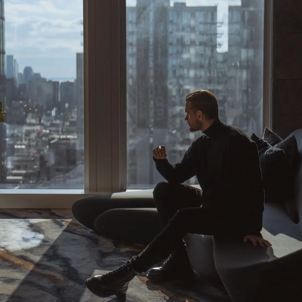 Person seated on curved couch in Equinox hotel suite, gazing at cityscape through floor-to-ceiling windows