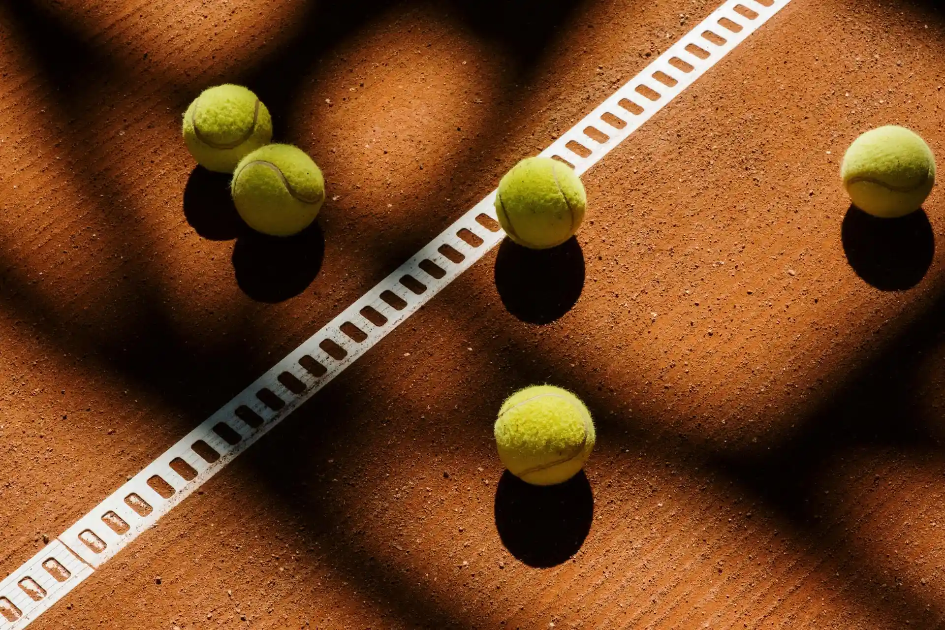Tennis balls scattered on a clay court under shadows near a white line