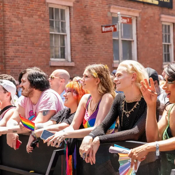 Colorful crowd with rainbow flags behind barricade at Pride event near Waverly Place street sign