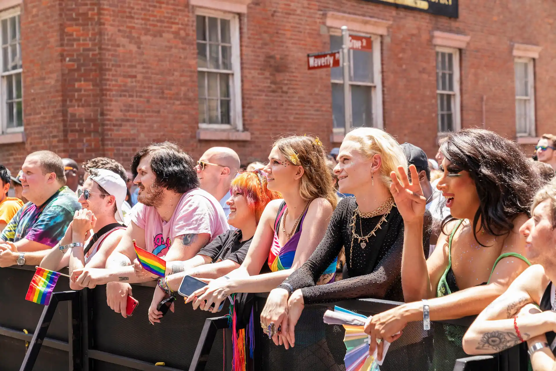 Colorful crowd with rainbow flags behind barricade at Pride event near Waverly Place street sign