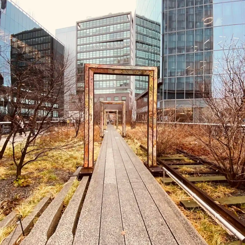 Elevated walkway with rust-colored arches and dry plants surrounded by glass buildings in urban park