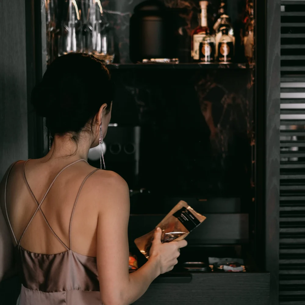 Person in satin dress examining minibar drawer with snacks and liquor bottles in marble-lined cabinet