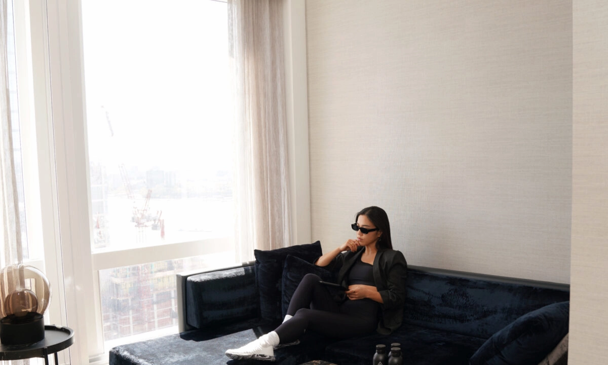 Person seated on blue velvet sofa with tablet and sunglasses in sunlit room overlooking urban construction