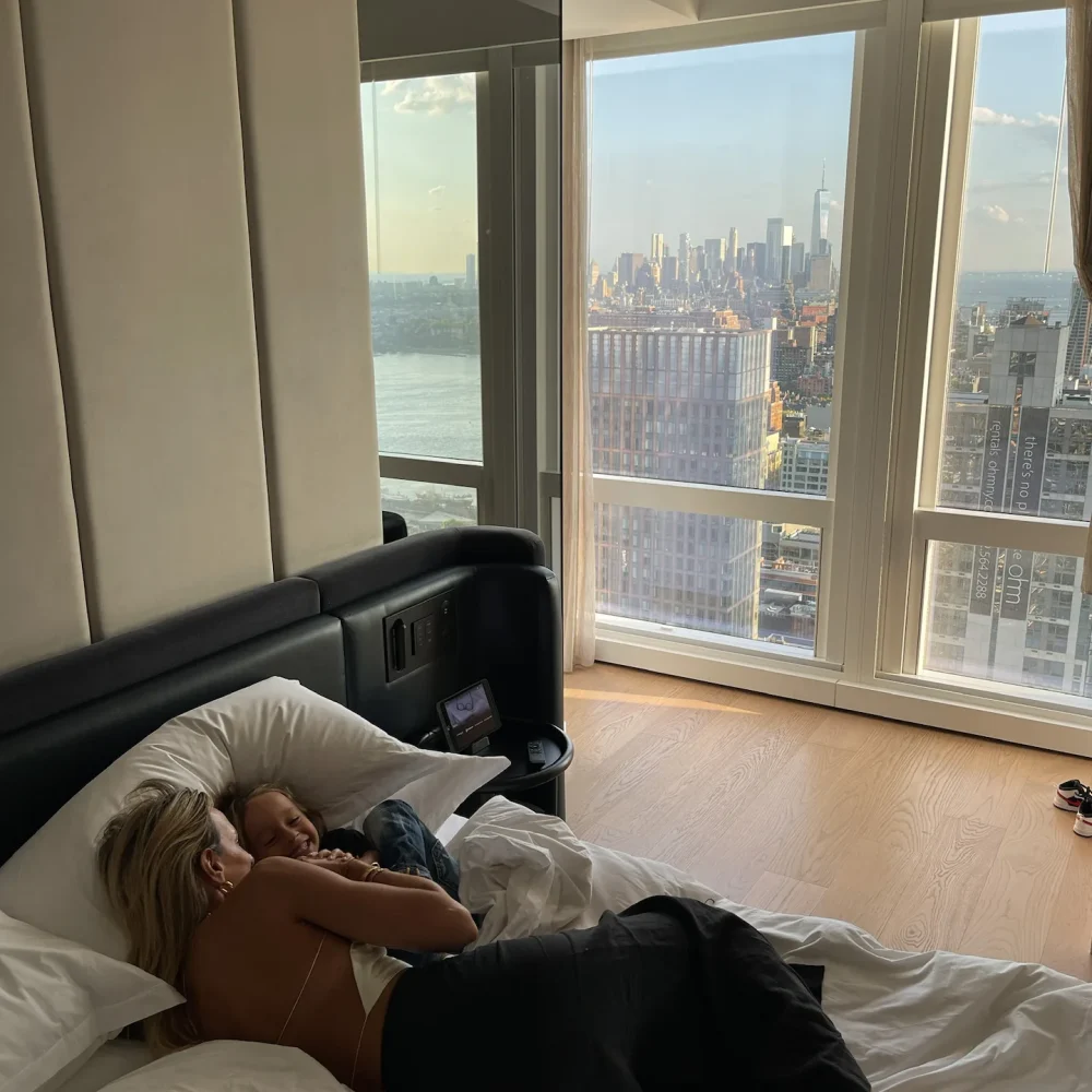 Woman cuddling with a child in a plush hotel bed, overlooking sweeping floor-to-ceiling views of the Manhattan skyline.