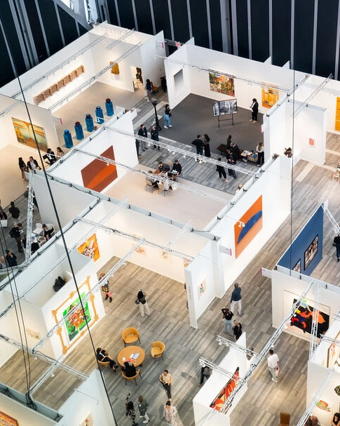 Overhead view of modern art fair with white booths, paintings, sculptures and visitors walking through