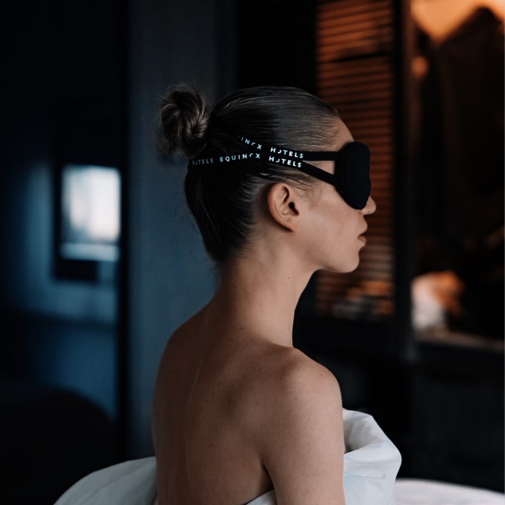 Woman sitting up in bed at Equinox Hotel, wearing a branded sleep mask and wrapped in white sheets, with only her shoulders and above visible as the rest of the room fades into a soft blur, highlighting a moment of restful stillness.