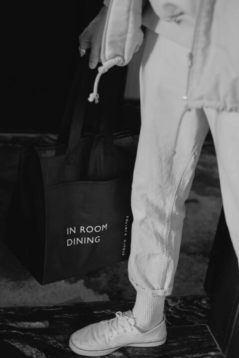 Person in white sneakers holding black “IN ROOM DINING” tote at Equinox, in elegant black-and-white hotel setting
