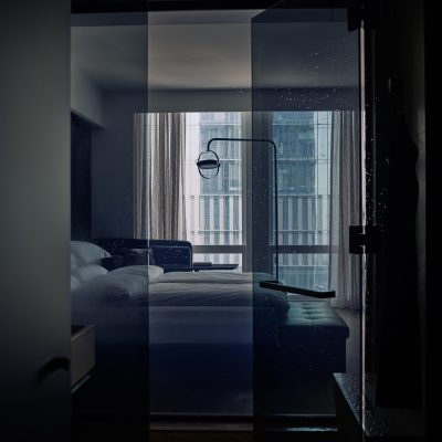 View of a sleek bedroom through wet glass, with white bed, bench, lamp, and city buildings outside window.