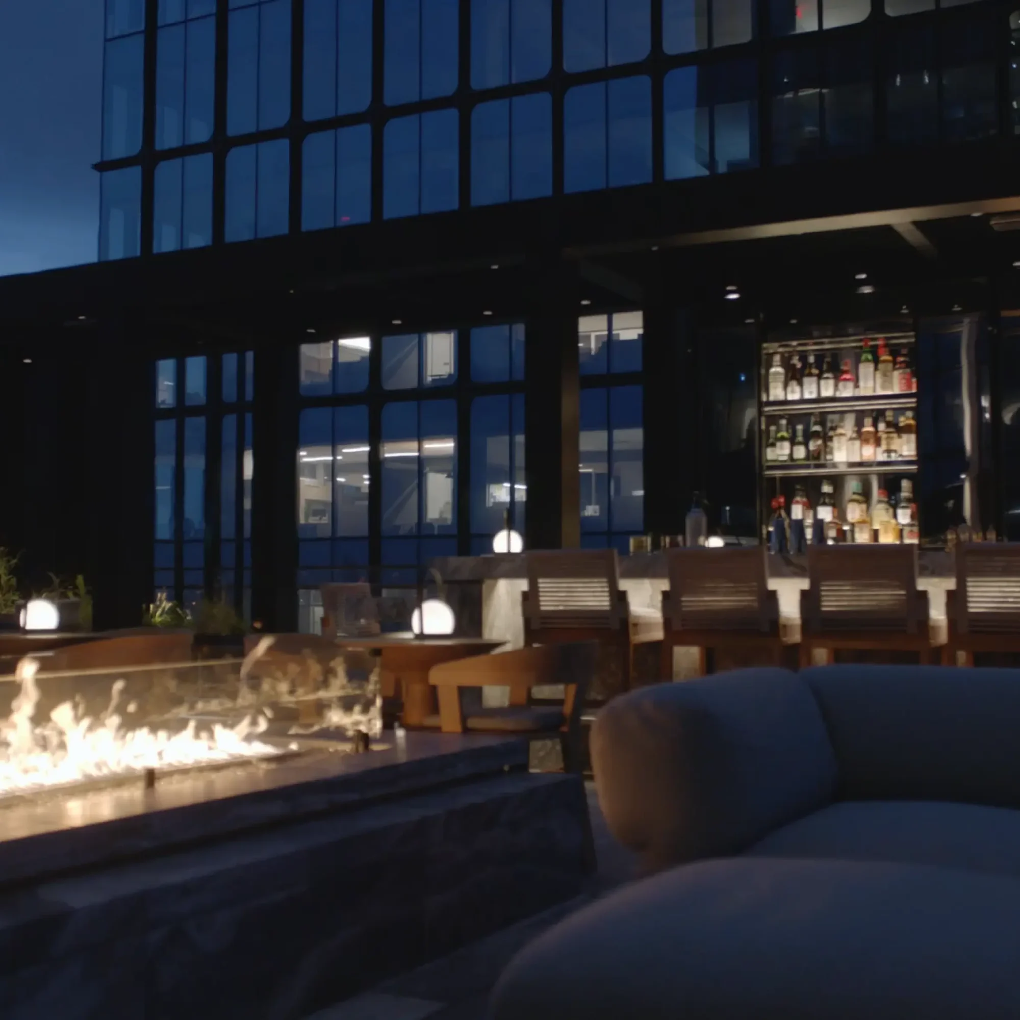 Rooftop lounge at night with fire pit, bar seating, and city skyline reflections