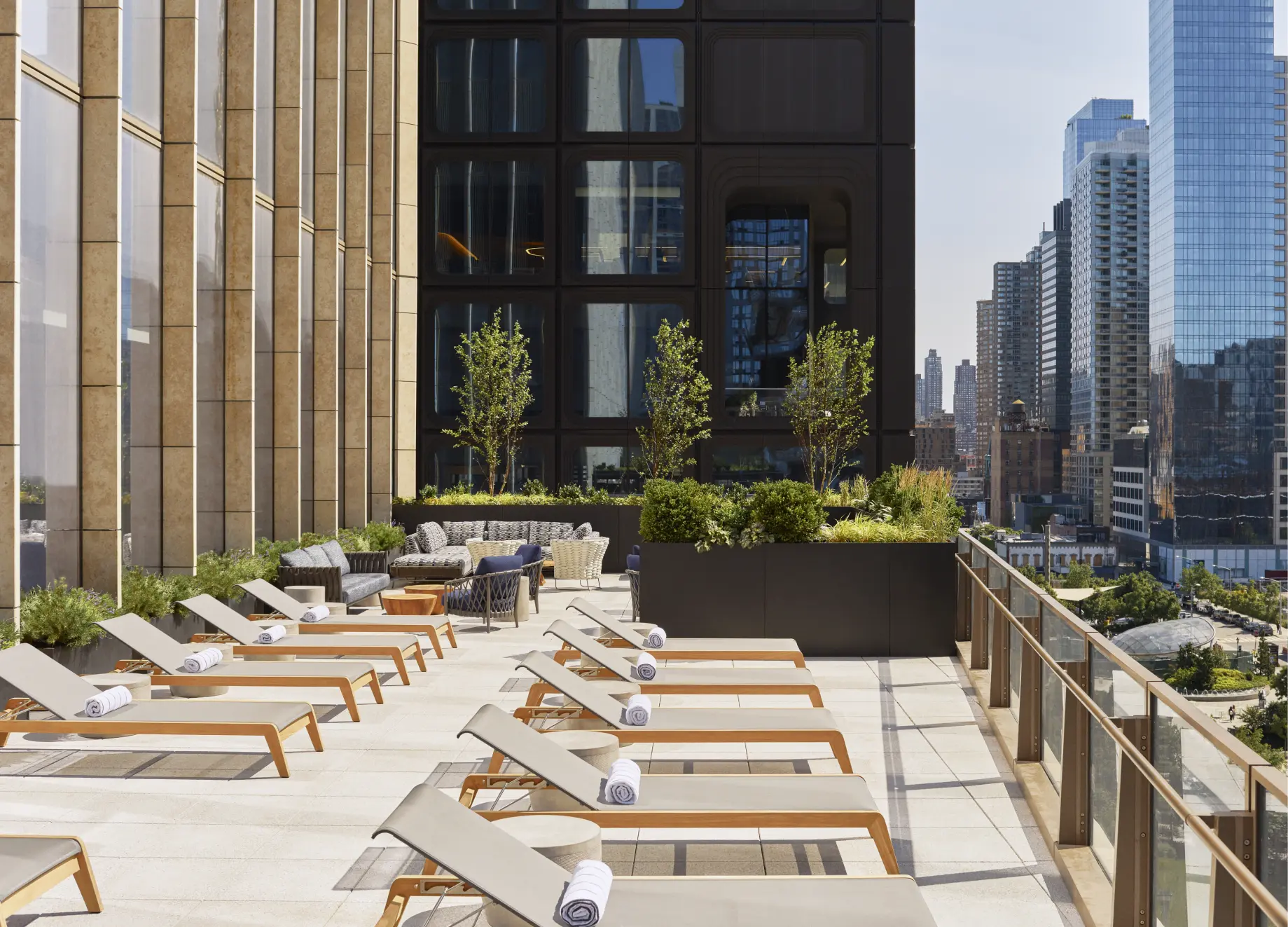 Rooftop terrace with cushioned lounge chairs, greenery, and city skyline views through glass railing