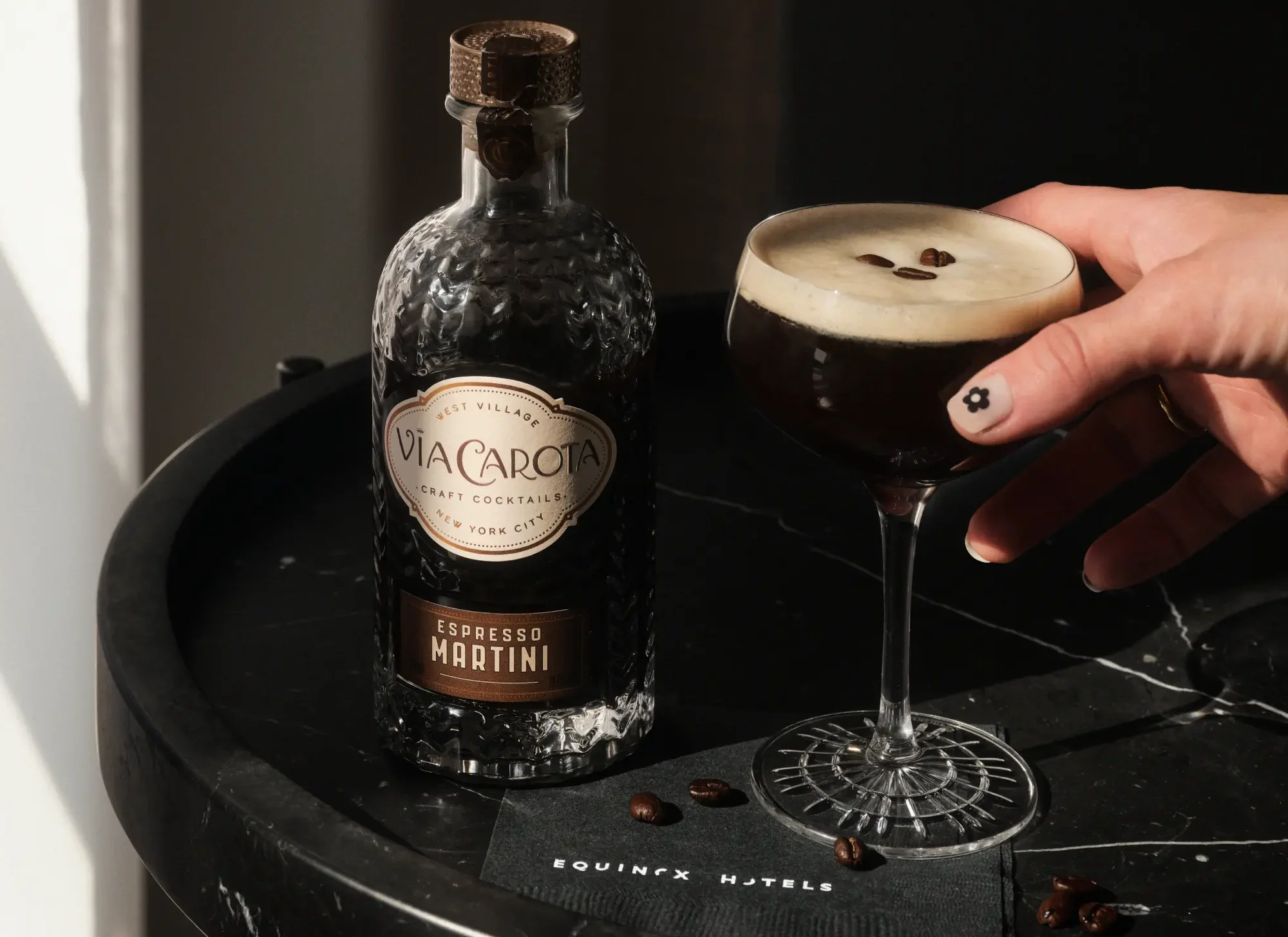 Hand holding espresso martini beside Via Carota bottle on marble table with Equinox napkin