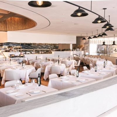 Estiatorio Milos interior with white tablecloths, pendant lights, open kitchen, and elegant curved ceiling
