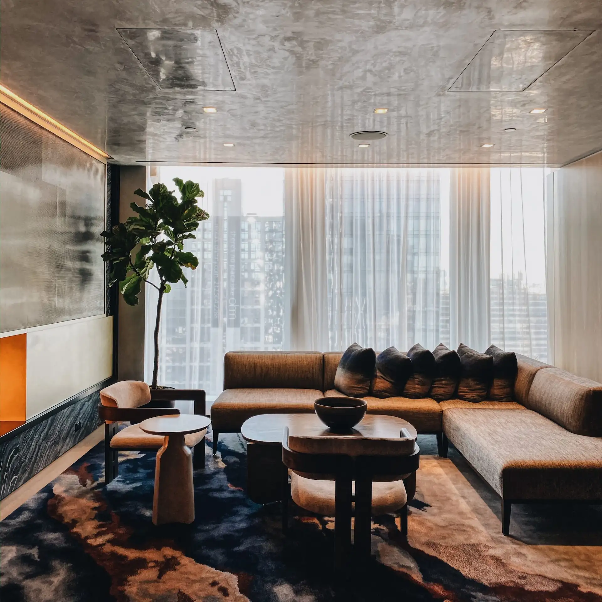 Modern living room at Equinox Hotels with brown sectional sofa, sculptural coffee tables, and city view through sheer curtains