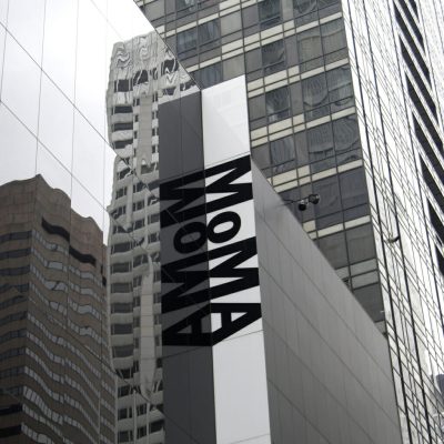 MoMA exterior with mirrored glass facade and bold vertical signage reflecting surrounding skyscrapers