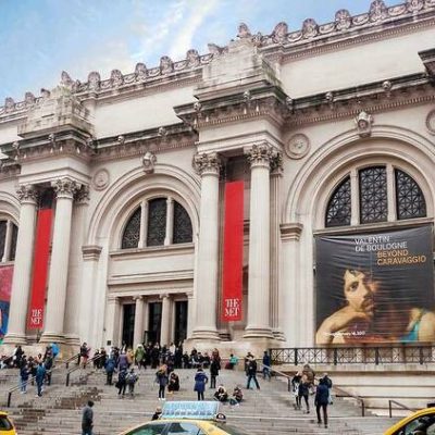 The Met facade with neoclassical columns, red banners, exhibition posters, and visitors on museum steps